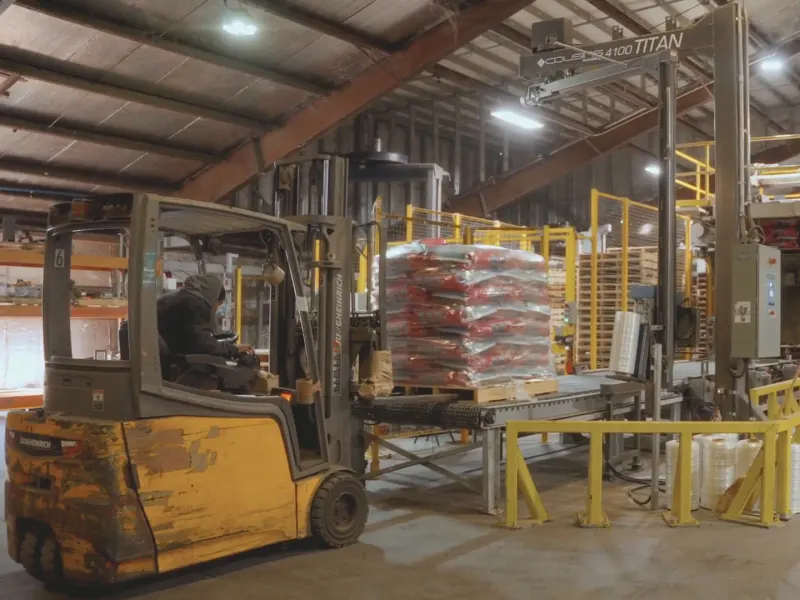 A forklift driver lifting a wrapped pallet of feed bags off the line